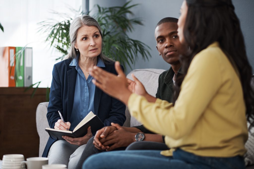 An older psychiatrist looks toward one of her patients in group therapy and takes notes.