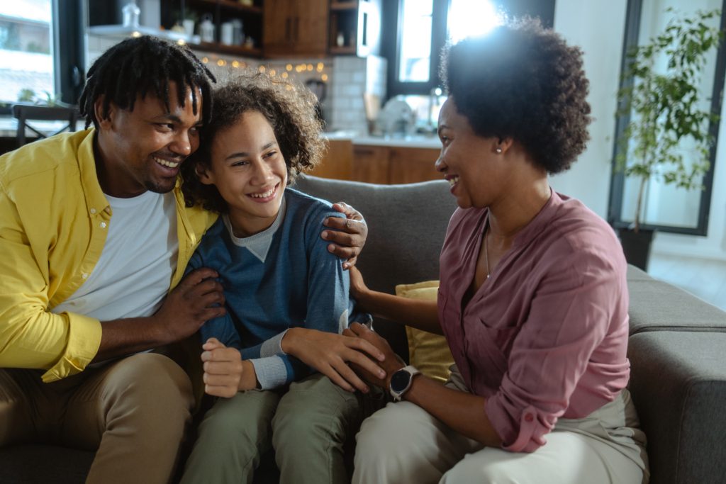 A mother and father smile while hugging their teenage daughter in their living room.